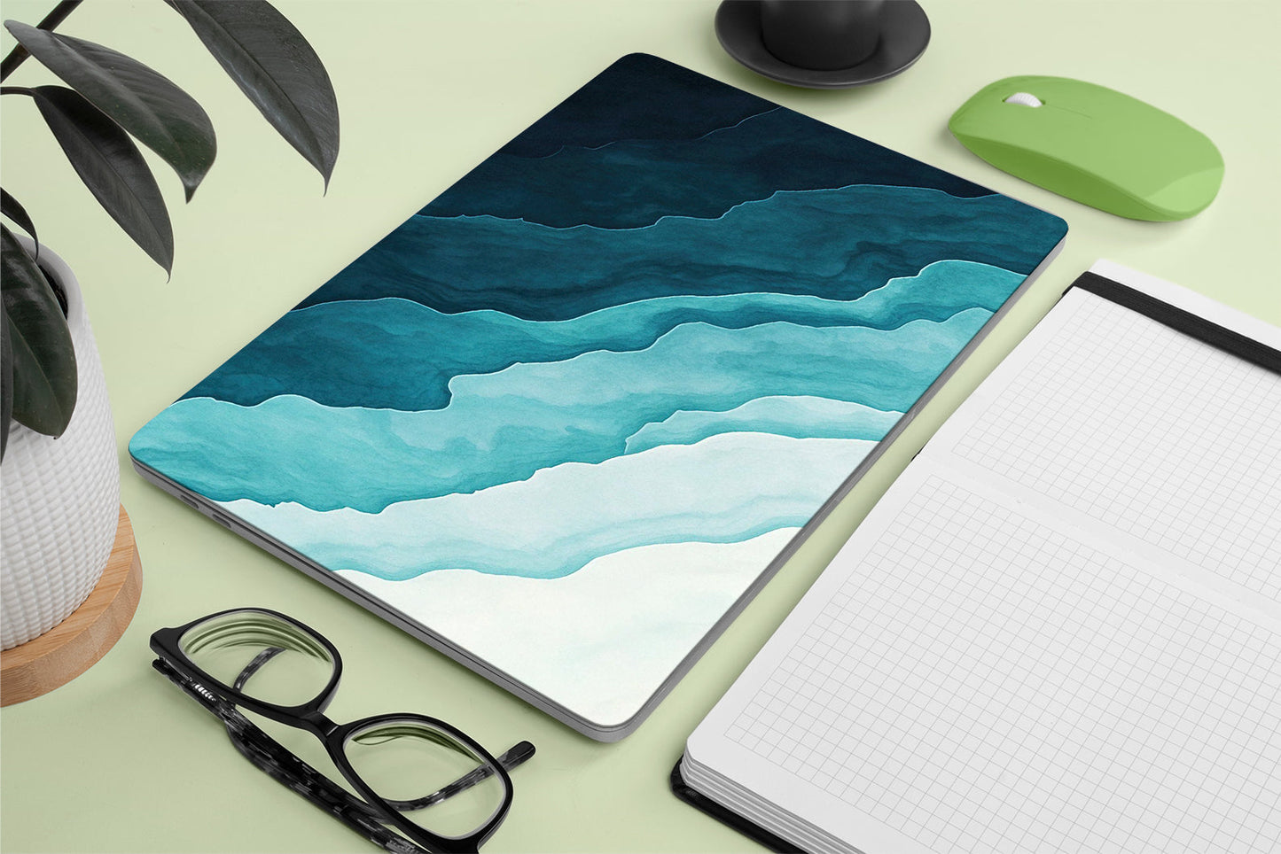 macbook with a blue and green abstract design on a light green surface with a notebook, glasses, and plant.